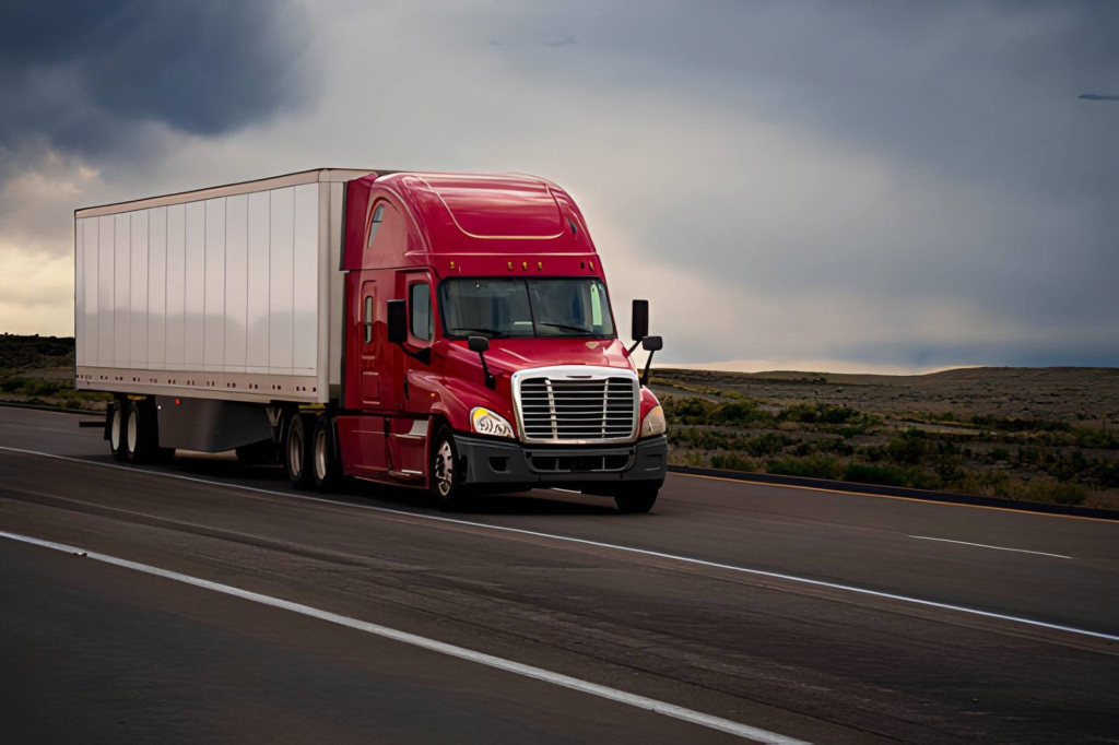 Red semi truck pulling a dry van trailer on a highway under stormy skies